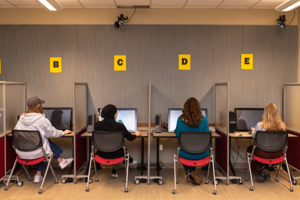 Four students modeling for labeled (A, B, C, D, E) computers in lab spaces.