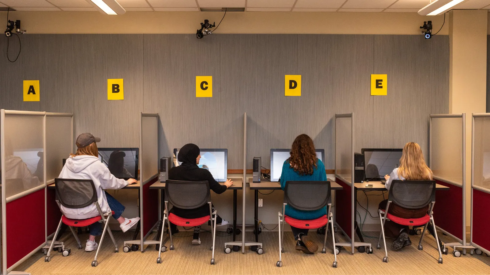 Four students modeling for labeled (A, B, C, D, E) computers in lab spaces.