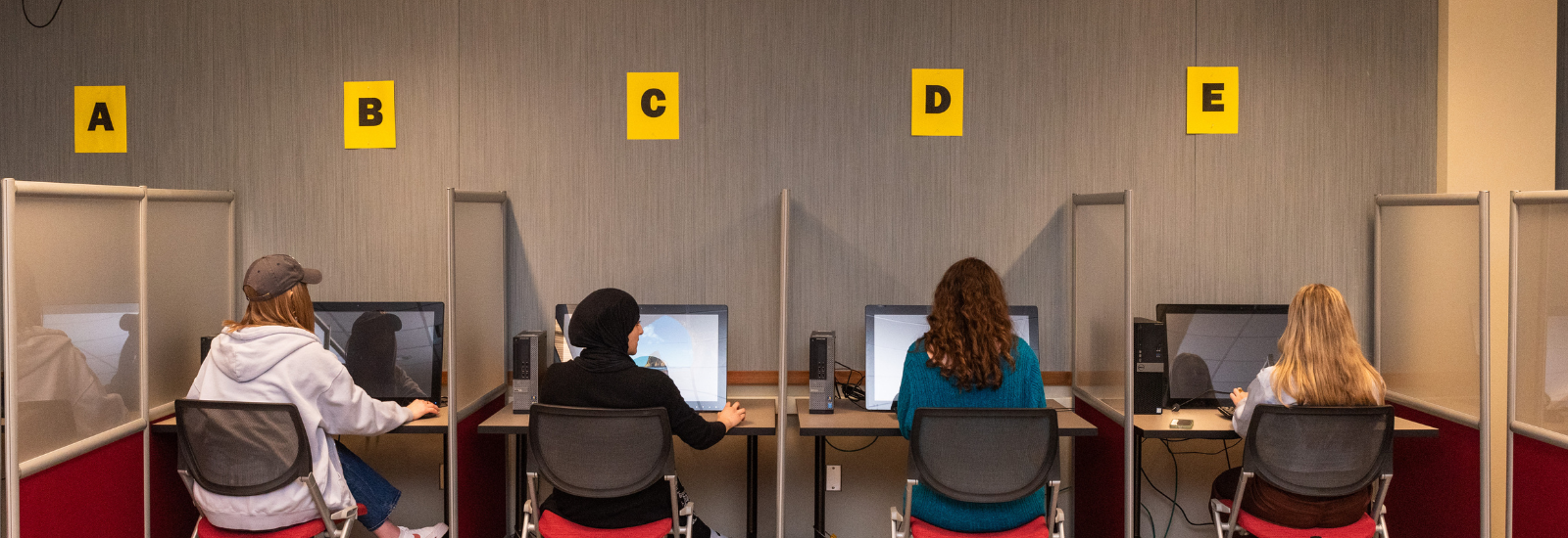 Four students modeling for labeled (A, B, C, D, E) computers in lab spaces.